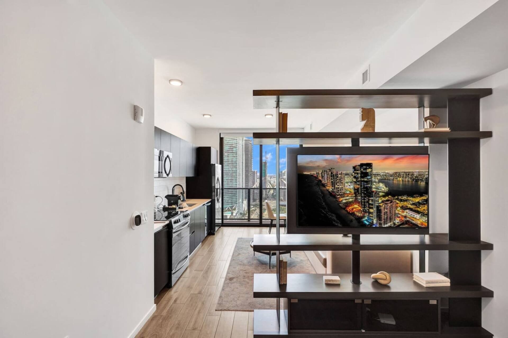 A modern living room with a flat-screen TV, a kitchen with grey cabinets, and a cityscape view through large windows.