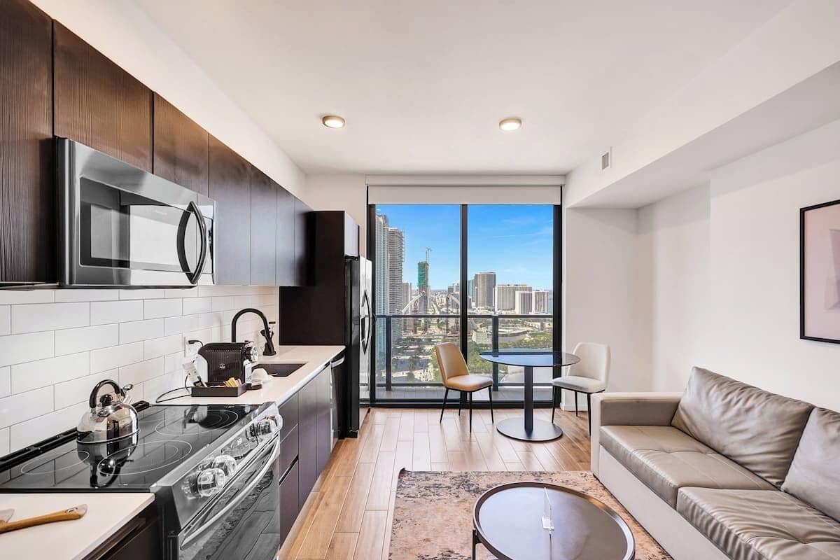 Una cocina moderna con estufa, microondas y hervidor, un comedor con mesa y sillas, y un amplio ventanal con vistas a la ciudad.