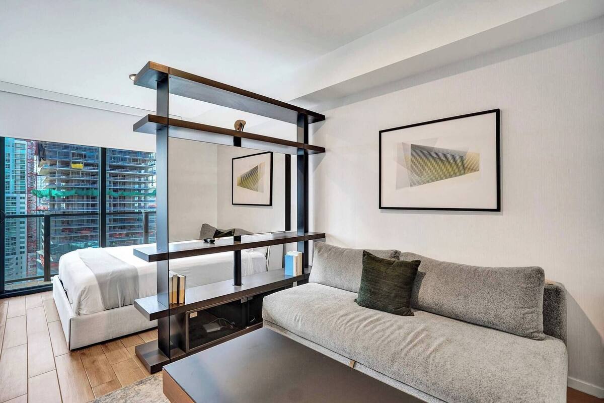 Una sala de estar moderna con sofá, mesa de centro, cama y un amplio ventanal con vista a un paisaje urbano.