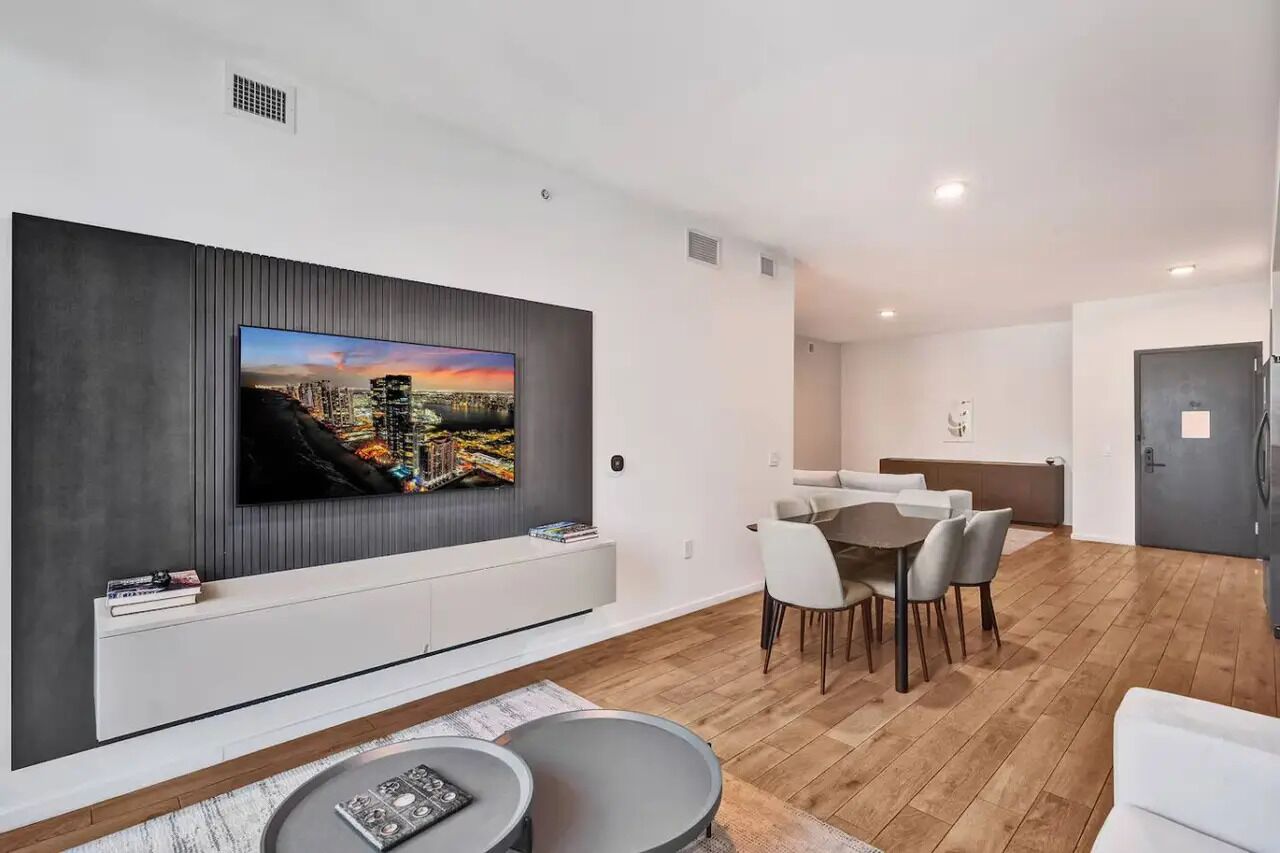 Ein modernes Wohnzimmer mit einem an der Wand montierten Flachbildfernseher, einem Essbereich mit weißem Tisch und Stühlen und einem Holzboden.