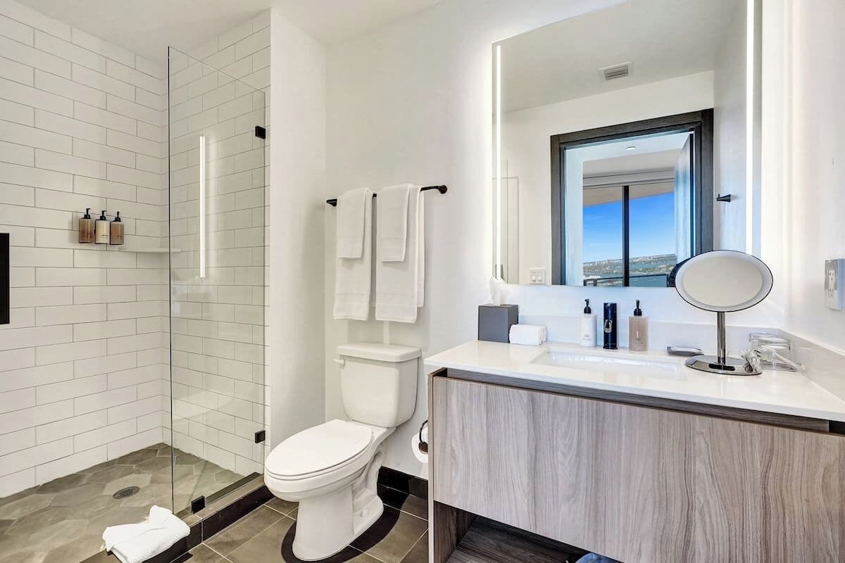 A modern bathroom with a glass-enclosed shower, white tiles, a toilet, a vanity with a mirror, and a view of the city through the window.
