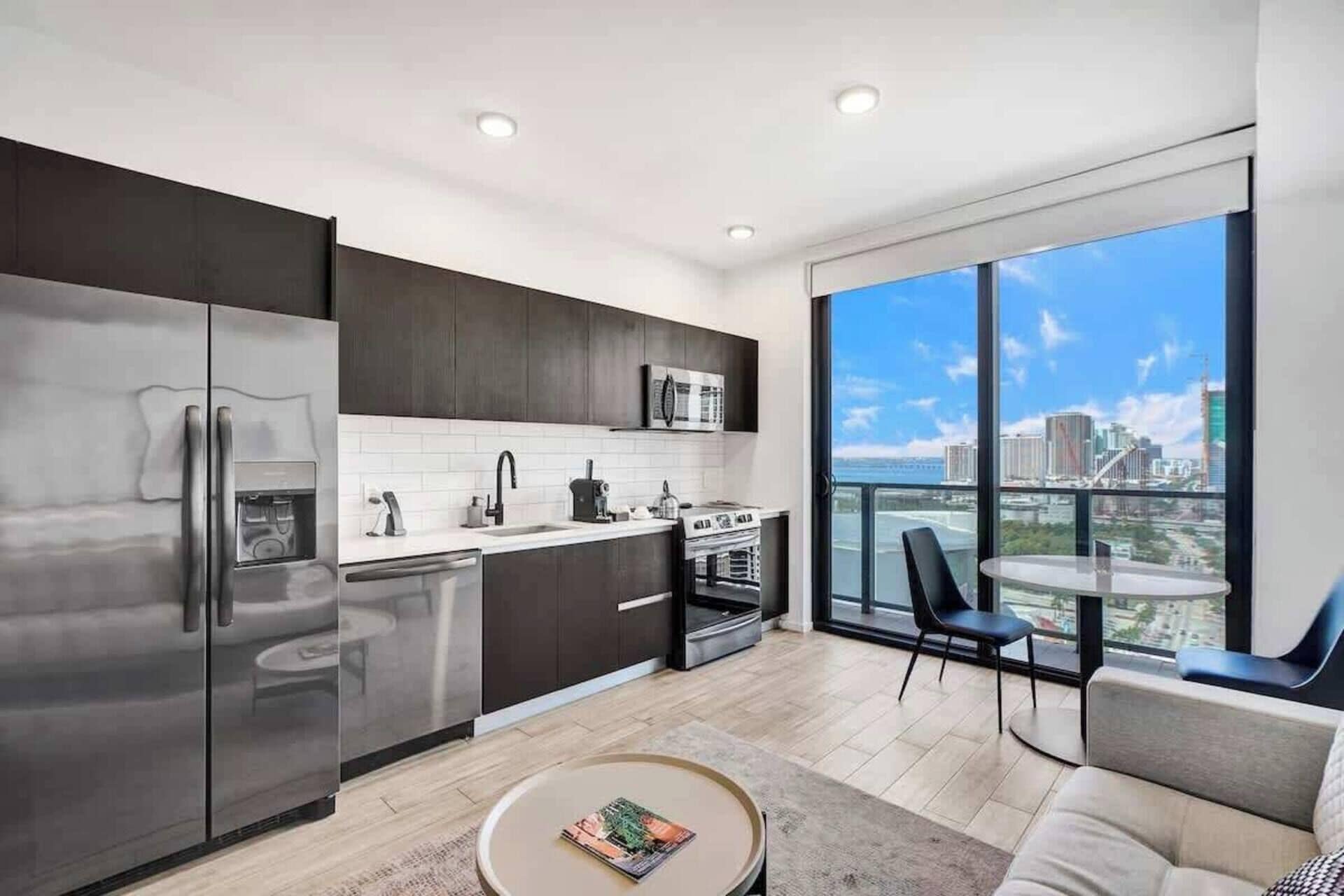 Una cocina moderna con un refrigerador grande, microondas y estufa, un comedor con una mesa redonda y sillas, y un ventanal con vista a la ciudad.