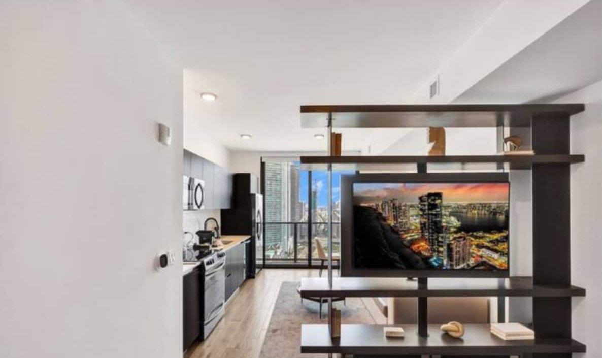 A modern living room with a large TV on a black stand, a kitchen in the background, and a view of the city from the window.