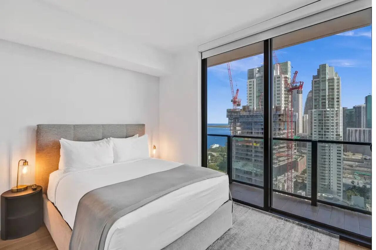 Una habitación de hotel moderna con una cama grande, una mesita de noche y vistas a un paisaje urbano con rascacielos.
