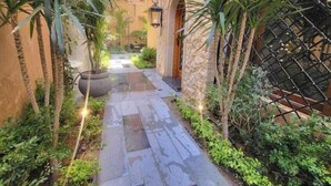 A narrow stone pathway flanked by lush greenery and a stone wall, leading to a building with an arched entrance. - 2BRs Garden side view-Three Sisters Villa 2 (New Cairo City)