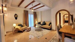 A spacious living room with a high ceiling, wooden beams, a large window with curtains, a sofa set, a coffee table, and a dining area with a table and chairs. - 6BRs with private entrance -Three Sisters Villa (New Cairo City)