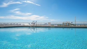 Una piscina azul cristalina con una escalera de metal, rodeada por una cerca blanca, y un cielo despejado al fondo.