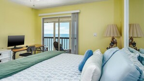 Una habitación con cama, escritorio con computadora, silla, lámpara y vista al mar a través de una puerta corrediza.