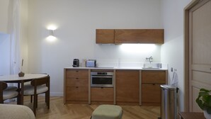 A compact kitchen with wooden cabinets, a built-in oven, a coffee maker, and a toaster. There is a small dining table with chairs and a trash bin.