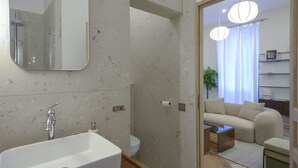 A modern bathroom with a large mirror, sink, and shower. A living area with a sofa and a coffee table is visible through an open doorway.