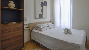 A bedroom with a wooden wardrobe, a bed with white linens, a large abstract painting, and a window with sheer curtains.