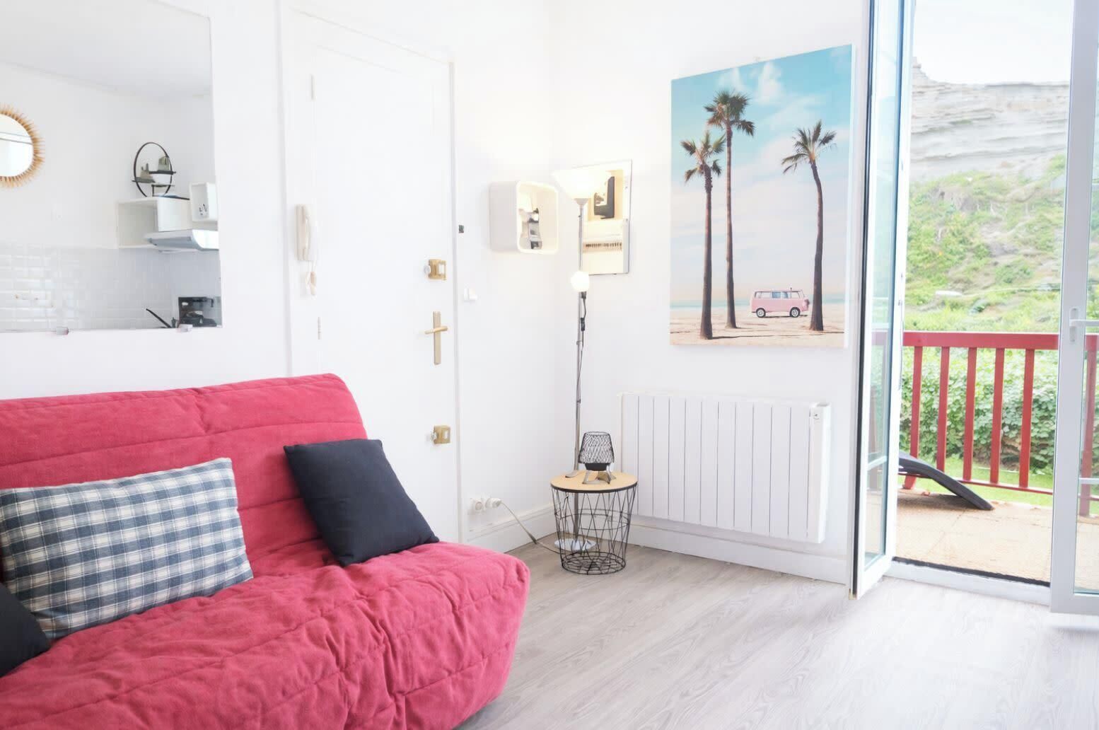 A room with a red sofa, a small side table, a radiator, a framed picture, and a balcony with a red railing.