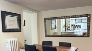 Ein Essbereich mit einem Holztisch und vier Stühlen, einem Heizkörper, einem gerahmten Bild und einem großen Spiegel, der eine Küche reflektiert.