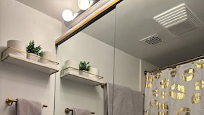 Ein Badezimmer mit einem weißen Waschbecken, goldenen Wasserhähnen und einem Spiegel darüber. Handtücher hängen an der Wand und ein Duschvorhang mit goldenem Muster ist angebracht.