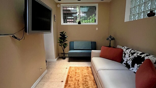 Ein Wohnzimmer mit Deckenventilator, einem flachbildfernseher an der Wand, einer Couch mit gemusterten Kissen, einem Fenster mit Blick auf Grünflächen und einem braunen Teppich.