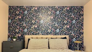Ein Schlafzimmer mit floralem Tapetenmuster, einem gepolsterten Kopfteil, einer gestreiften Bettdecke, einem Nachttisch mit Lampe und einer dunkelbraunen Kommode.