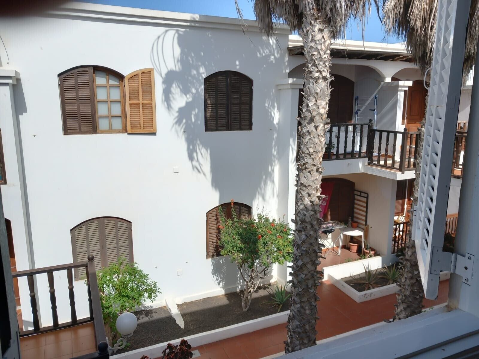 A white building with multiple balconies, a courtyard with plants, and a palm tree.