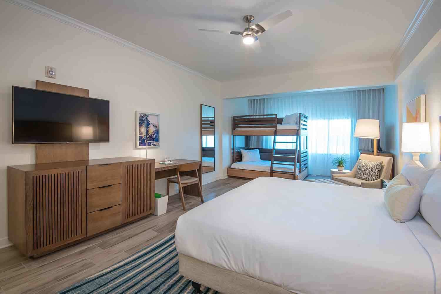 A hotel room with a large bed, a desk with a chair, a TV, bunk beds, a ceiling fan, and a window with curtains.