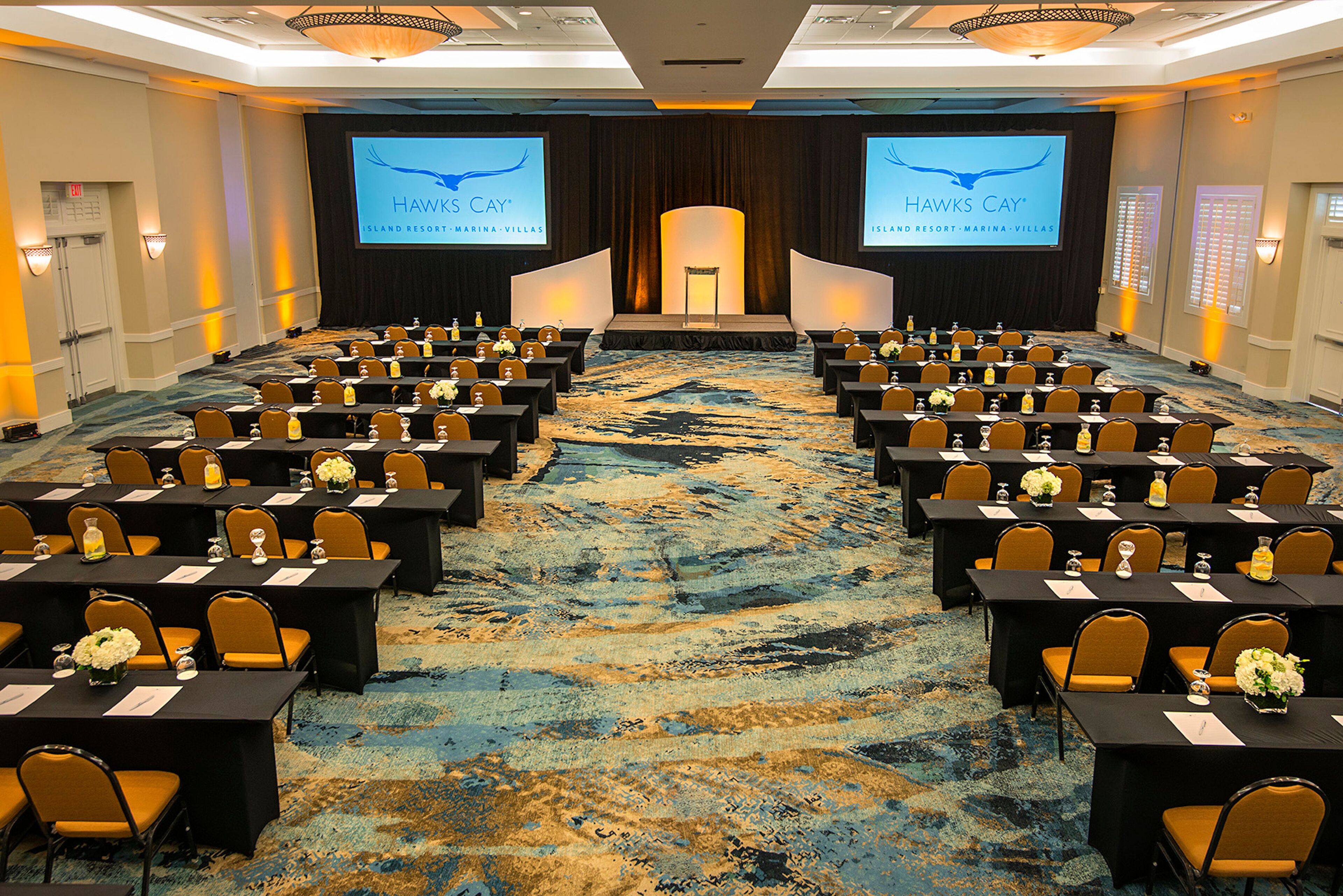 A conference room with rows of tables and chairs, a stage with a screen displaying 'Hawks Cay' branding, and a carpeted floor.