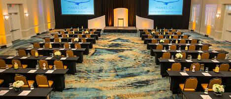 A conference room with rows of tables and chairs, a stage with a screen displaying 'Hawks Cay' branding, and a carpeted floor.