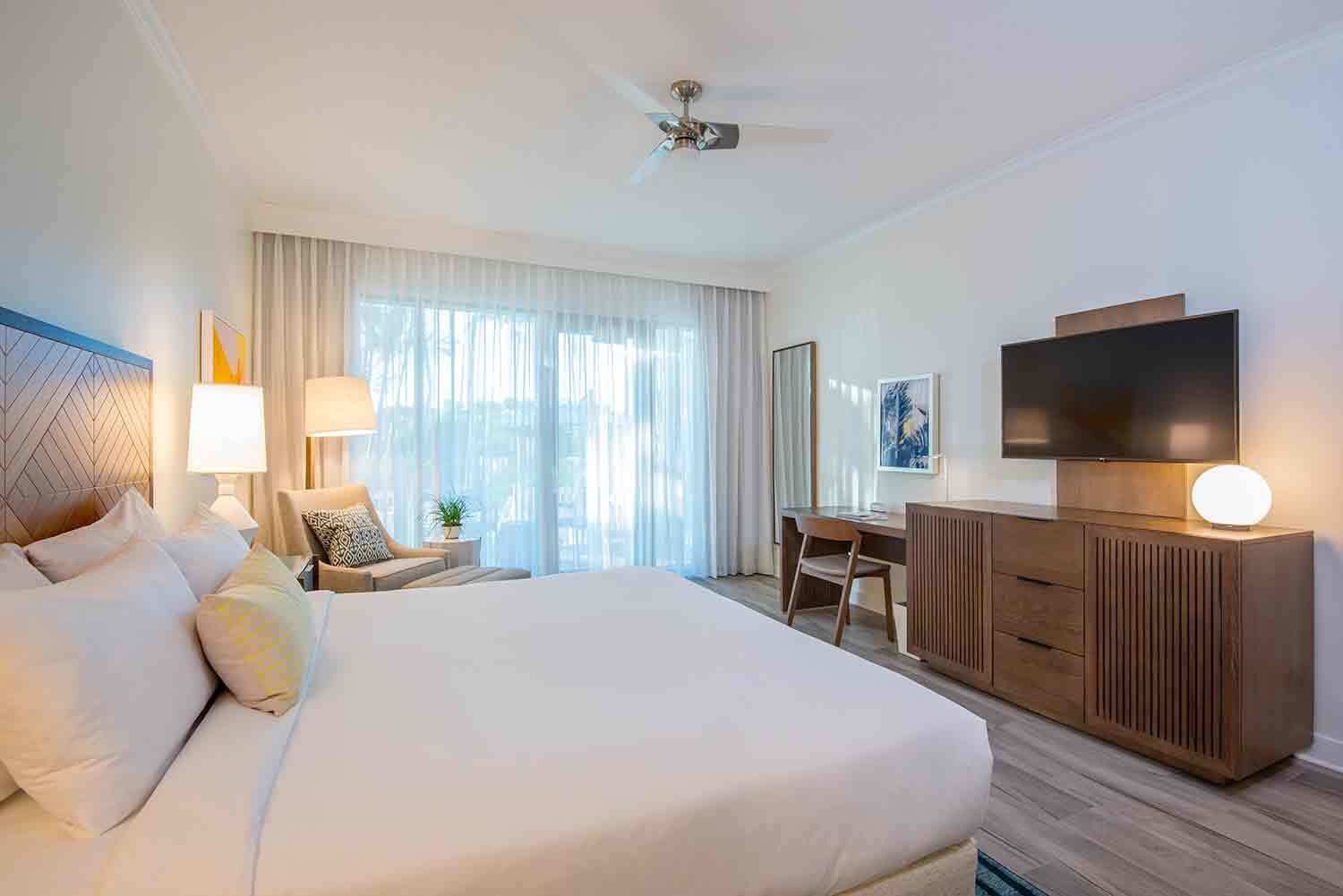 Une chambre d’hôtel avec un grand lit, un bureau, une chaise, une télévision et un ventilateur de plafond.