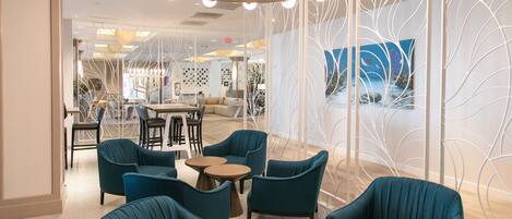 A modern lounge area with teal chairs and round tables, a decorative white partition, and a ceiling light fixture.