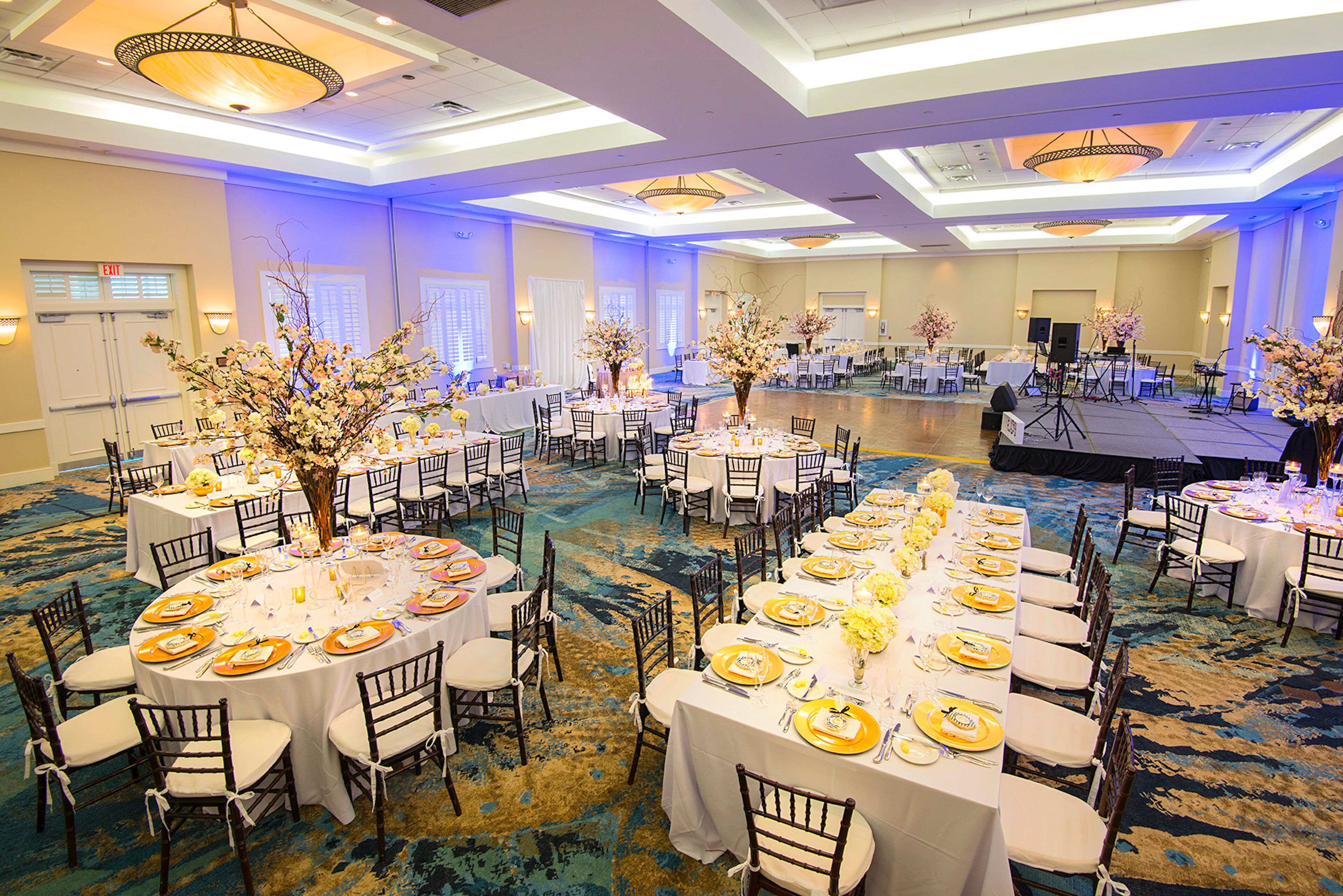 Uma sala de banquetes com mesas redondas preparadas para um evento formal, com arranjos florais no centro e um palco ao fundo.