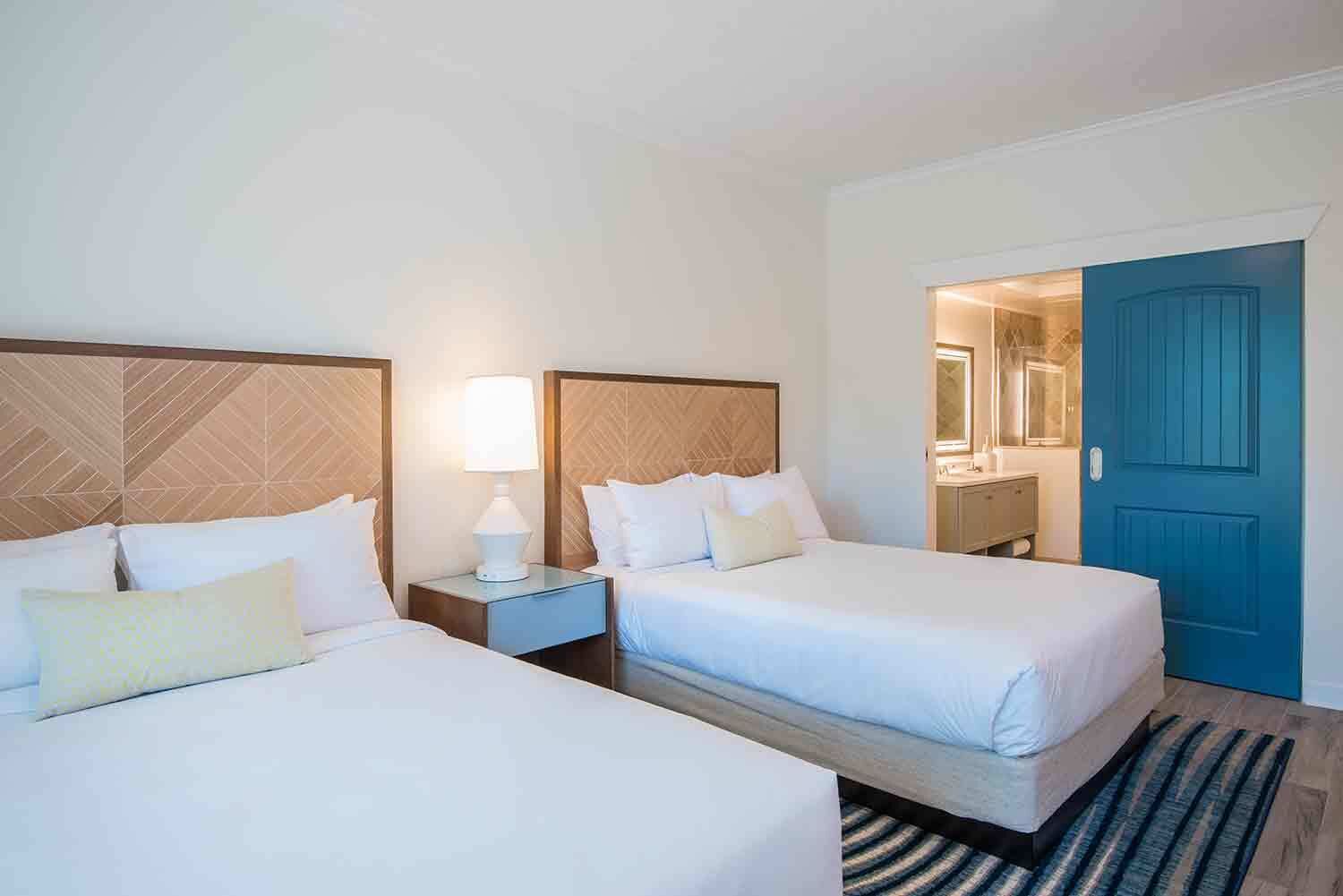 A hotel room with two beds, a nightstand with a lamp, a blue door, and a mirror.