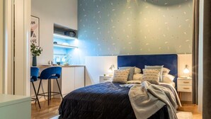 A bedroom with a dark blue headboard, a bed with multiple pillows, a bedside table with a lamp, and a small desk area with a chair. - Black & White Studio downtown- M2 metro 4-6 tram (Budapest)