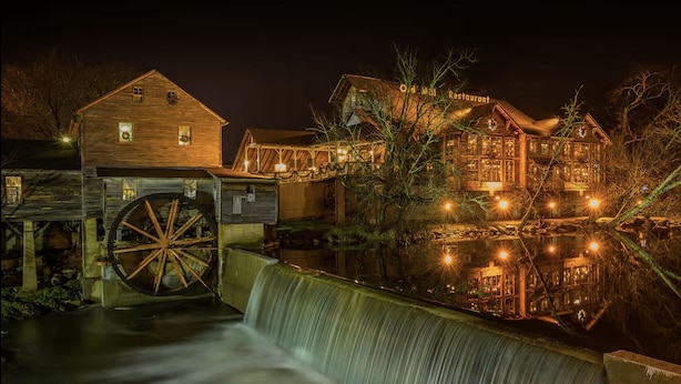 Ein historisches Gebäude mit einem großen Wasserrad, einer Holzkonstruktion mit dem Schild "Old Mill Restaurant" und einem Spiegelbild im Wasser.