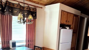 Een compacte keuken met houten kastjes, een witte koelkast, een eettafel met stoelen en een hangende lamp.