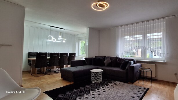 Una sala de estar moderna con un sofá modular, un comedor con mesa y sillas blancas, y un gran ventanal con persianas.