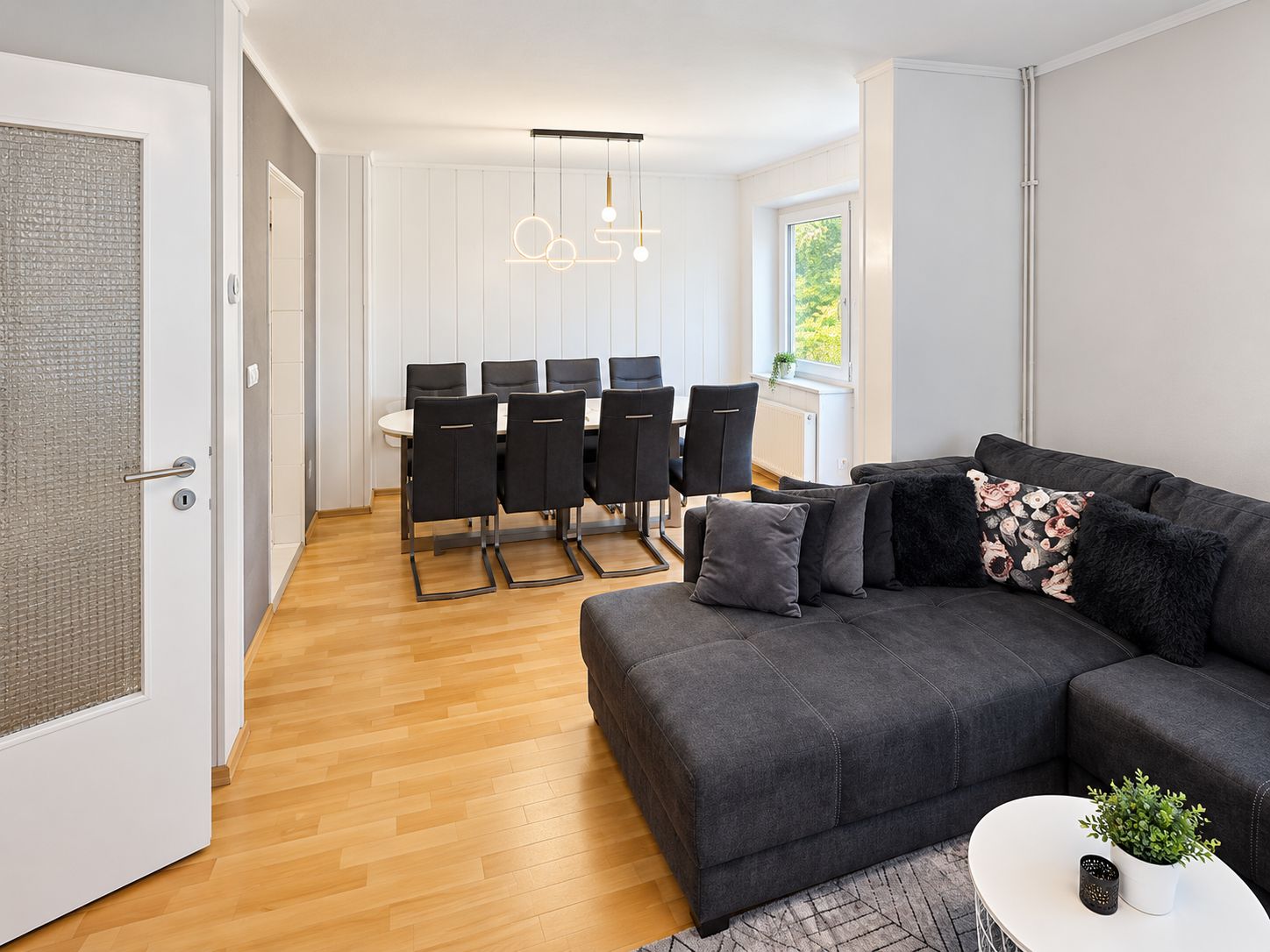 A modern living room with a dark grey sofa, a white coffee table, and a dining area with a white table and black chairs.