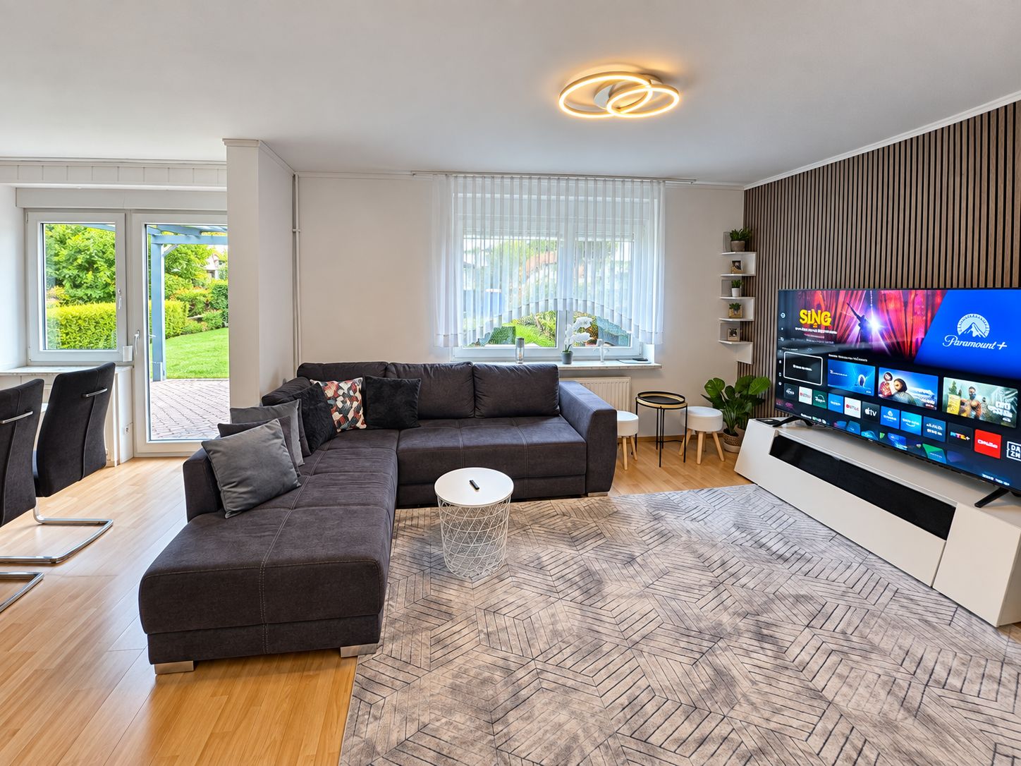 A modern living room with a sectional sofa, a coffee table, a TV mounted on the wall, and a view of the outdoors through large windows.