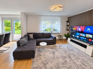 Een moderne woonkamer met een hoekbank, een salontafel, een aan de muur bevestigde televisie en uitzicht op het groen door grote ramen.