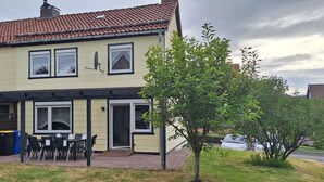 Une maison à deux étages avec un patio couvert, un espace barbecue et un petit arbre dans le jardin.
