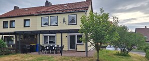 Ein zweistöckiges Haus mit überdachter Terrasse, Grillplatz und einem kleinen Baum im Vorgarten.