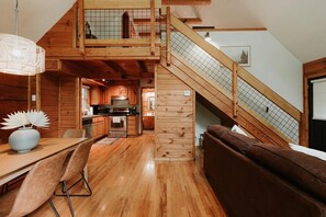 Um amplo quarto em estilo loft com escada de madeira, área de jantar com mesa e cadeiras, e um espaço de estar aconchegante com sofá marrom.