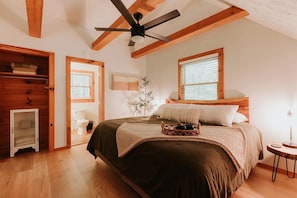 Quarto com cama grande, vigas de madeira, ventilador de teto, janela com persianas e mesa de cabeceira.