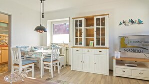 A dining area with a wooden table and chairs, a white cabinet with glass doors, and a television on a wooden stand. - Vacation apartment Möwenkoje (Norddeich)