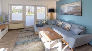 A modern living room with a grey sofa, a wooden coffee table, and a large window with blinds. - Vacation apartment Möwenkoje (Norddeich)