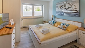 A double bed with yellow and white bedding, a wooden nightstand, a lamp, and a painting of a sailboat on the wall. - Vacation apartment Möwenkoje (Norddeich)