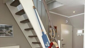Una escalera con una escoba azul apoyada, un flotador rojo y blanco, y una silla azul en la parte inferior.