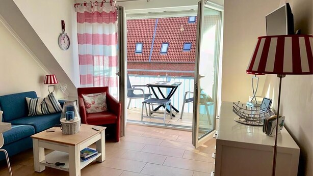 Un salón con sofá azul, una silla roja, una mesa de centro y un ventanal de vidrio que da a un balcón con un edificio de tejado rojo.