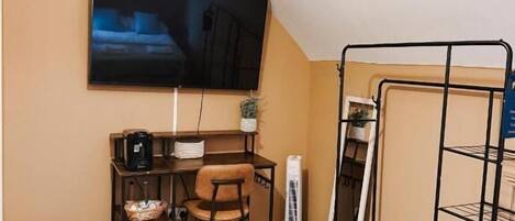 Quarto pequeno com uma cama, uma escrivaninha com cadeira, uma televisão fixada na parede e uma estante de metal.