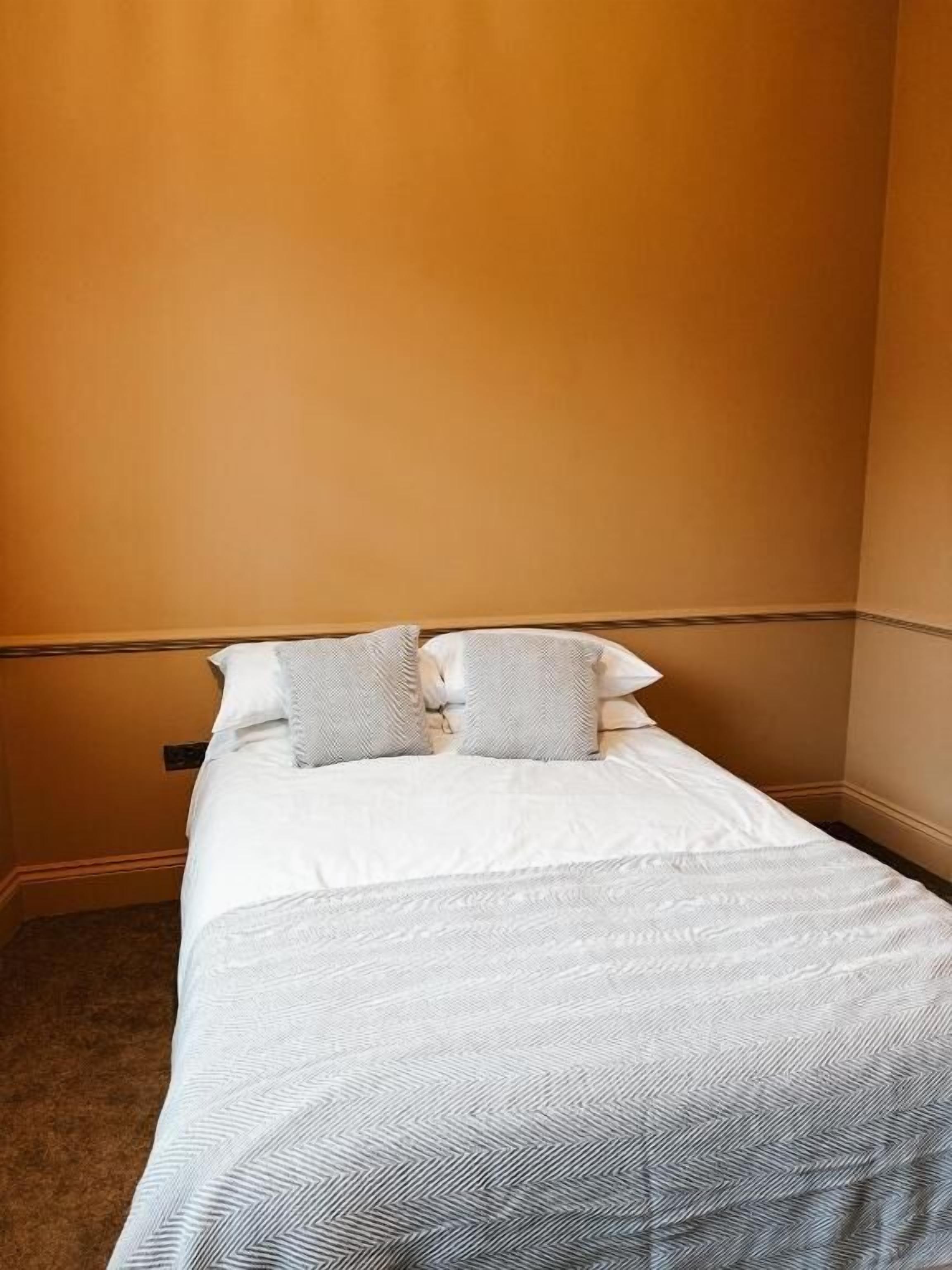 Una cama individual con una colcha blanca y gris estampada, dos almohadas y un cabecero de madera en una habitación con paredes anaranjadas.