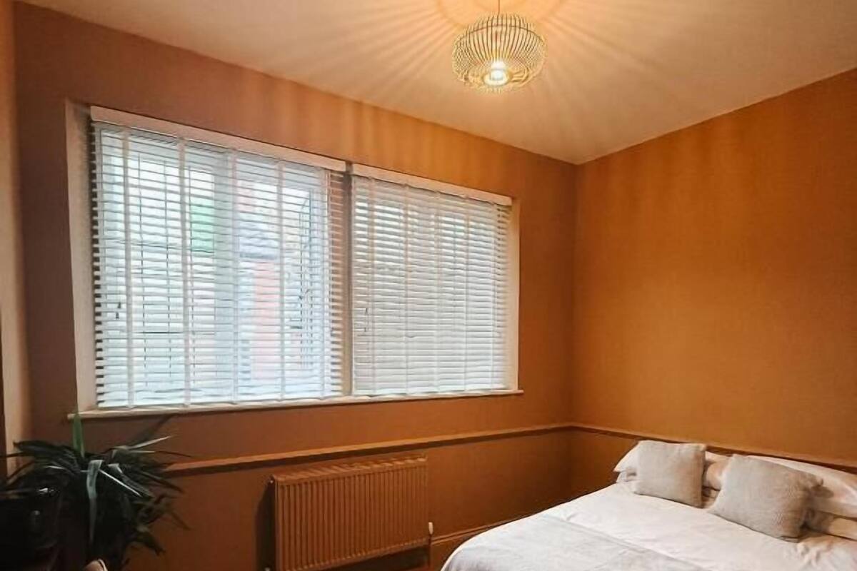 A bedroom with a bed, a radiator, a window with blinds, and a plant.
