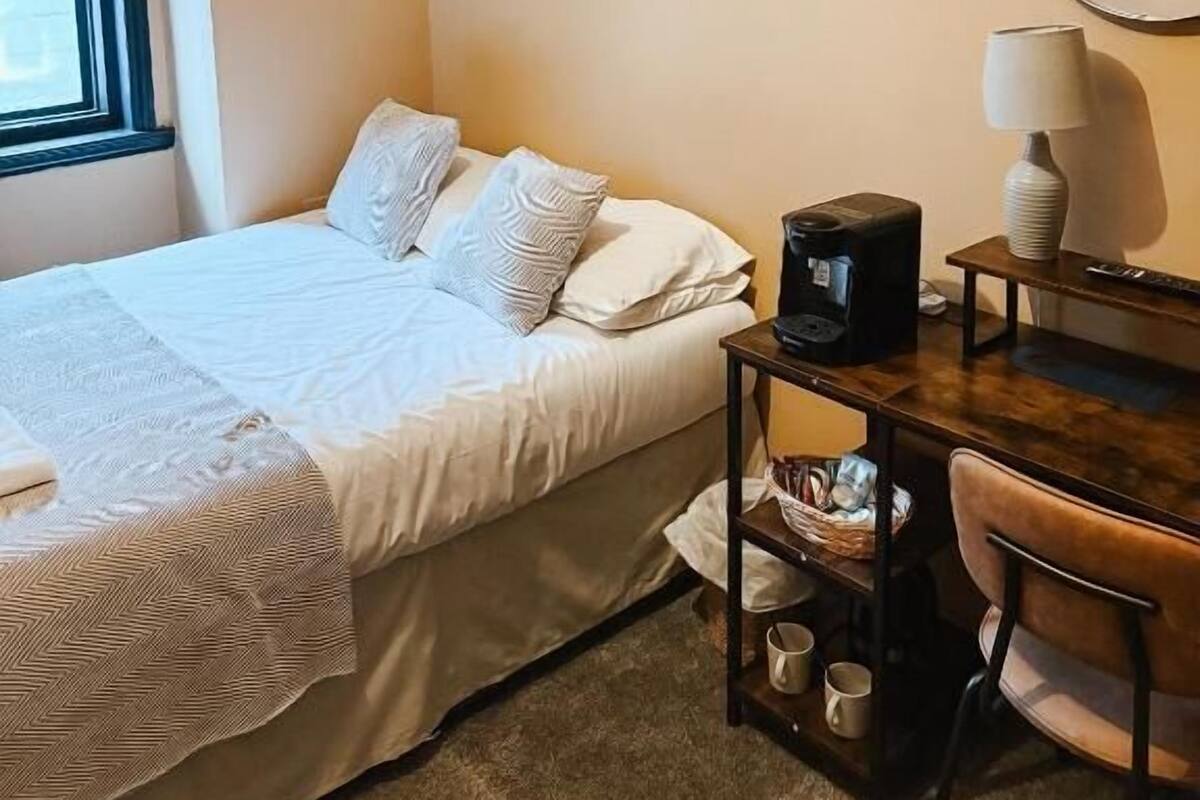 A hotel room with a bed, a desk with a lamp, a coffee maker, and a chair.