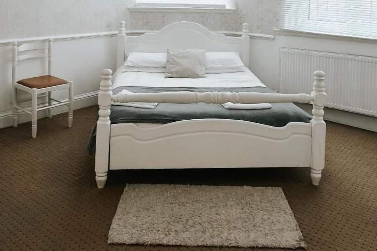 A white four-poster bed with a wooden chair, a white radiator, and a window with blinds.