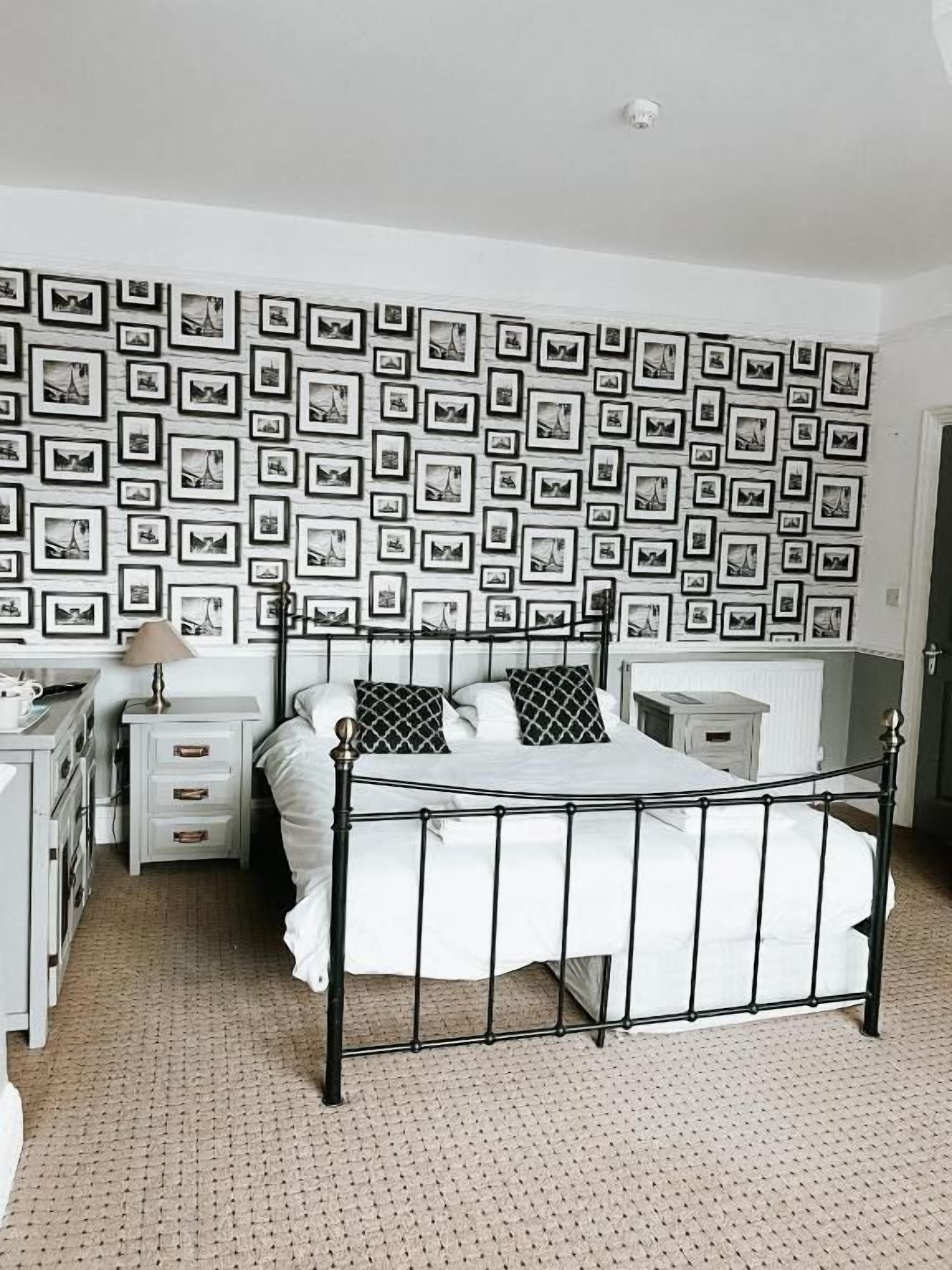 A bedroom with a metal bed frame, white bedding, a dresser, a nightstand, and a wall covered with framed pictures.